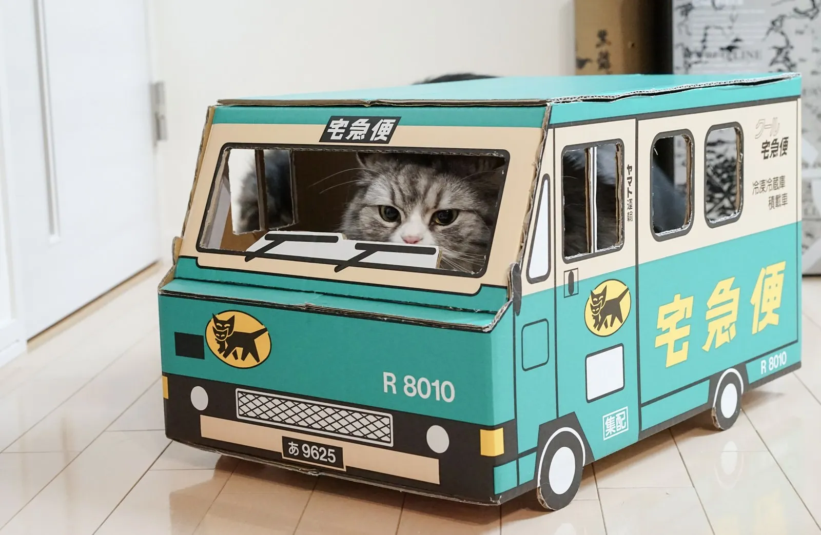 A cat peeking out of the driver's window of a cardboard box shaped like a delivery truck