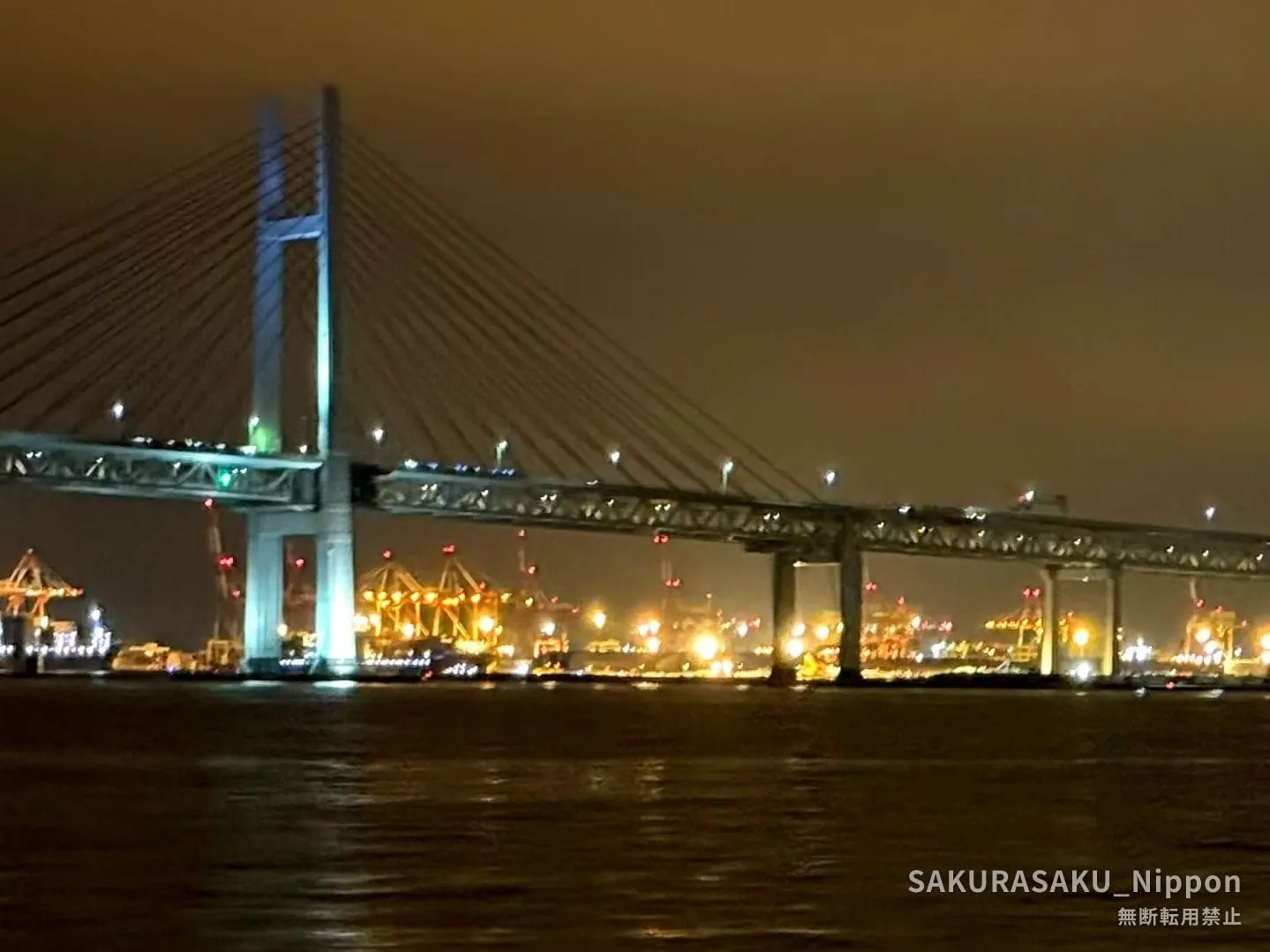 The Yokohama Bay Bridge lit at night, seen from the water, with the lights of the container terminal beyond